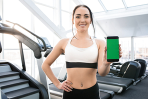 Waist up portrait of fit young woman wearing sports clothes presenting mobile app holding smartphone with green screen and smiling at camera