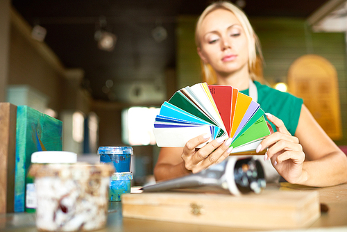 Portrait of pretty blonde woman looking at color swatches choosing palette for art and craft project in workshop, focus on foreground