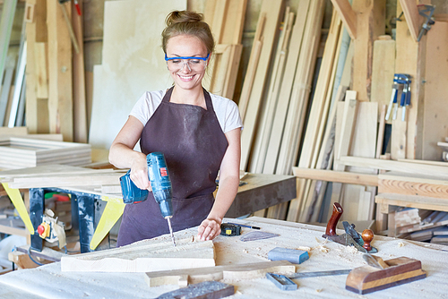Portrait of cheerful young woman working in carpenters shop drilling holes in piece of wood while making furniture, copy space