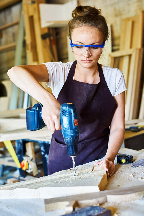 Portrait of young woman working in carpenters shop drilling holes in piece of wood while making furniture