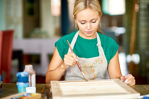 Front view portrait of pretty blonde woman enjoying work in art studio painting frame with silver paint, making DIY interior decoration