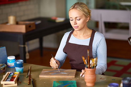 Portrait of pretty blonde woman enjoying work in art studio painting picture on wooden base making DIY decor