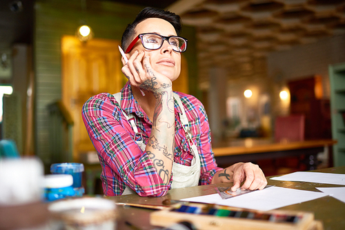 Portrait of creative young woman with tattoos on her arms sitting at table in artists workshop looking away pensively looking for inspiration
