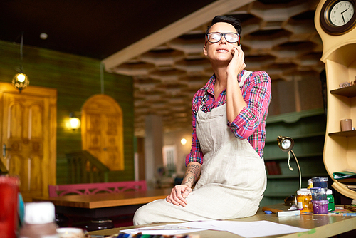 Portrait of modern female artist wearing apron taking break from work sitting on table and speaking by phone in workshop