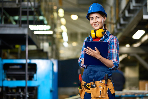 Portrait of female construction worker working in modern plant , smiling filling in papers on clipboard