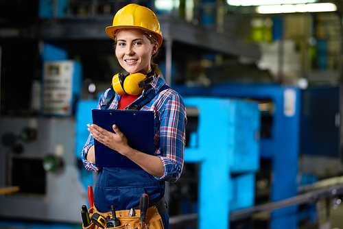 Waist-up portrait of attractive young technician in checked shirt and overall  with wide smile while carrying out inspection at production department of plant, blurred background