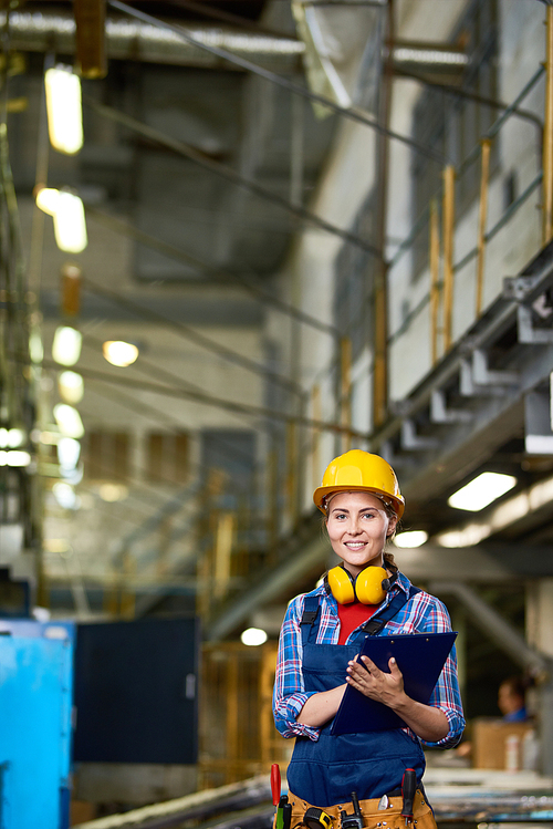 Portrait of young woman in hardhat working in modern plant, smiling at camera holding clipboard
