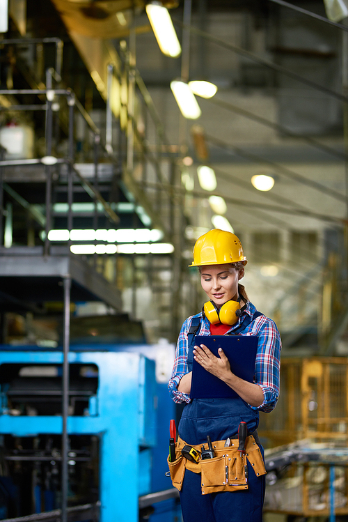 Portrait of pretty young woman wearing  hardhat looking clipboard writing something while  working in modern plant