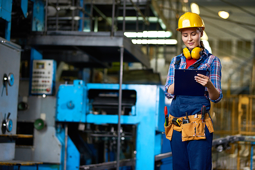 Confident young worker in protective helmet taking necessary notes while inspecting industrial machine, portrait shot