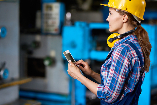 Side view portrait of female machine operator using digital tablet while working with machines in modern factory