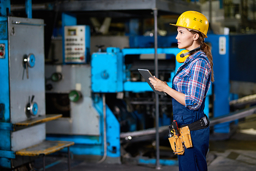 Side view portrait of female machine operator using digital tablet while working with power units in modern factory