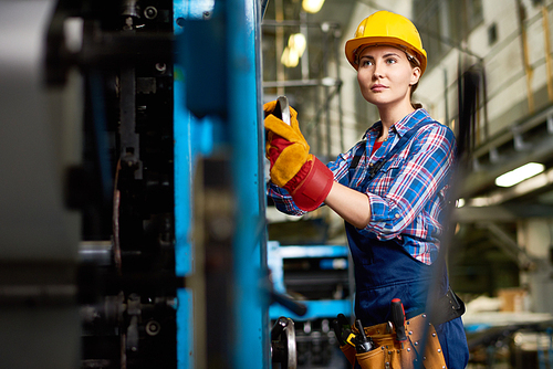 Confident young engineer wearing overall and checked shirt looking away while carrying out inspection at manufacturing plant, blurred background