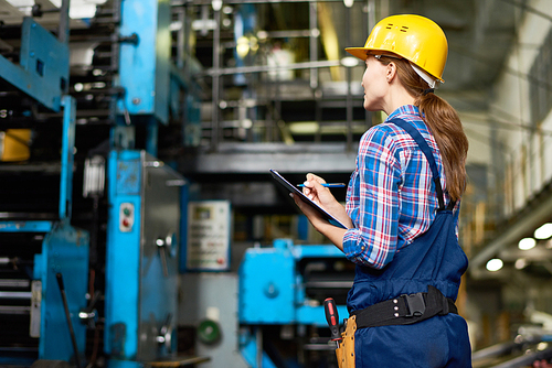 Portrait of female factory worker writing on clipboard  while checking operating machines in modern plant