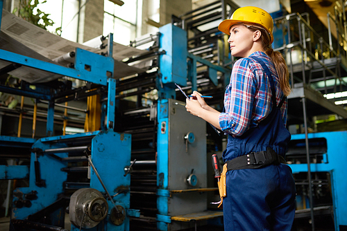 Back view of confident young worker wearing overall and checked shirt taking notes in clipboard while taking inventory at manufacturing plant
