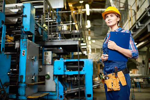 Portrait of young woman working in modern factory, walking between operating machines holding clipboard