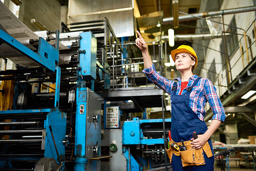 Portrait of talented factory worker in overall and checked shirt pointing at something while working at production department