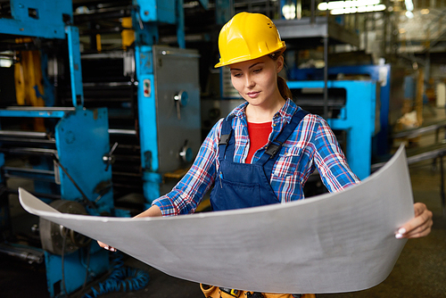 Attractive young industrial engineer wearing protective helmet standing at production department of modern plant and studying blueprint
