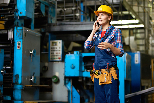Portrait of young woman working in modern factory speaking by phone between rows of machines, copy space