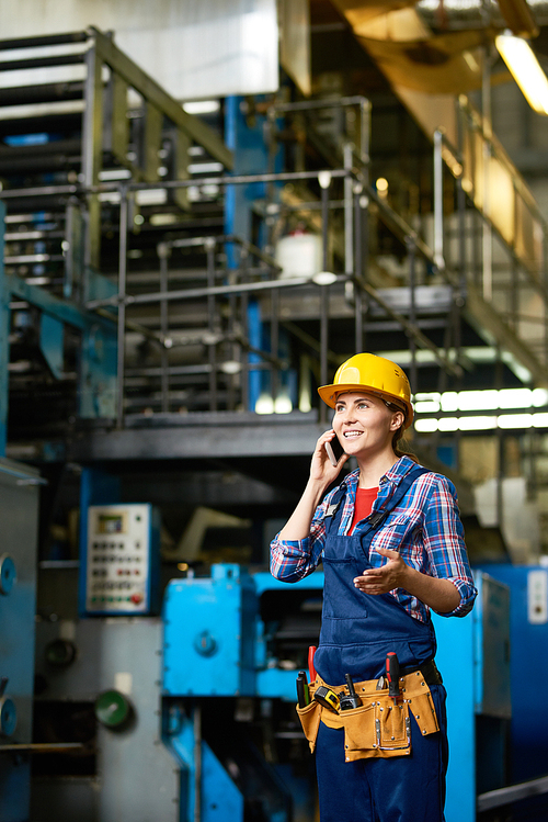 Portrait of young woman working in modern factory speaking by phone between rows of machines and smiling