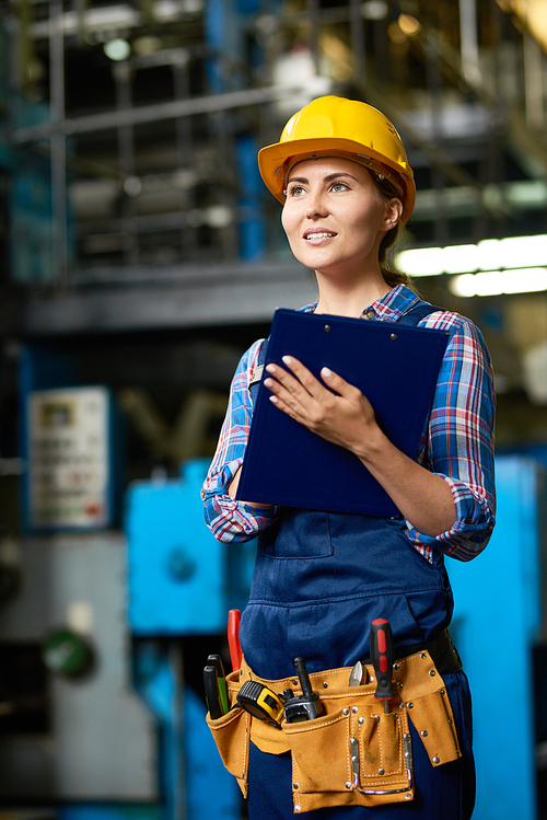Portrait of female factory worker wearing hardhat holding clipboard and smiling posing in modern workshop