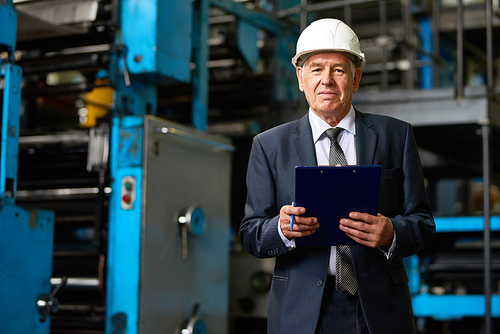 Portrait of senior factory owner  wearing hardhat on tour in modern factory  and smiling holding clipboard