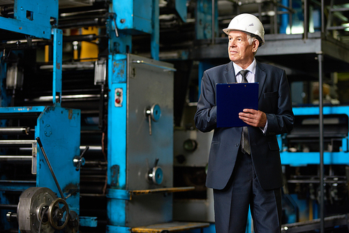 Senior investor in suit and protective helmet taking necessary notes in clipboard while walking along production department of modern plant, portrait shot