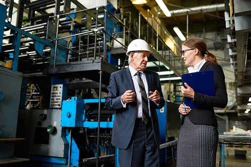 Portrait of senior businessman wearing hardhat talking to assistant manager in modern factory workshop, copy space