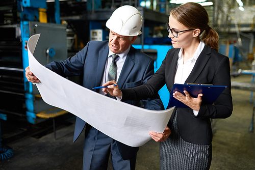 Portrait of senior businessman looking at floor plans of modern factory with young assistant manager explaining the construction