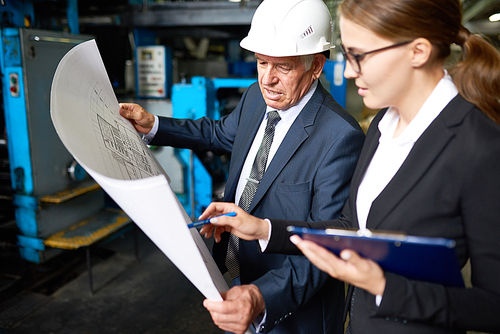 Portrait of senior businessman looking at floor plans of modern factory with young assistant manager in workshop