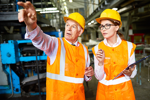 Portrait of senior factory worker explaining something to young woman pointing away at machines,  both wearing hardhats and reflective jackets