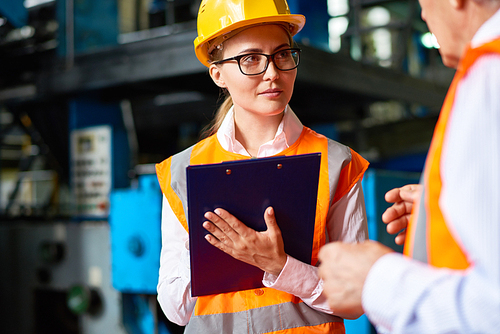 Portrait of young woman making notes on clipboard while talking to senior worker in  modern workshop,  both wearing hardhats and reflective jackets