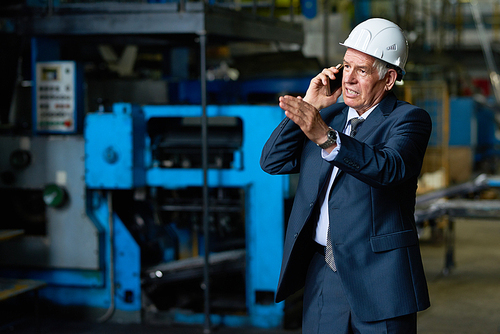 Confident senior entrepreneur wearing suit and hardhat talking to his colleague on smartphone while walking along manufacturing plant