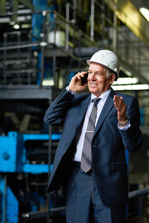 Portrait of senior businessman wearing hardhat speaking by phone and smiling in workshop of modern factory