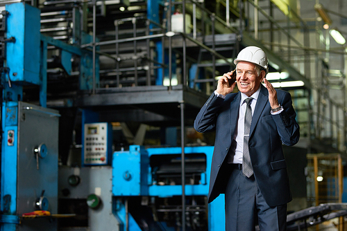 Portrait of senior businessman wearing hardhat speaking by phone and smiling in workshop of modern factory, copy space