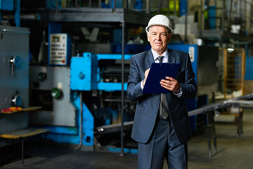 Portrait of smiling senior businessman wearing hardhat  holding clipboard in workshop of modern factory