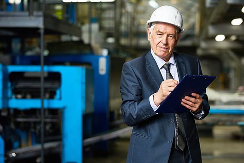 Portrait of senior businessman wearing hardhat  holding clipboard in workshop of modern factory