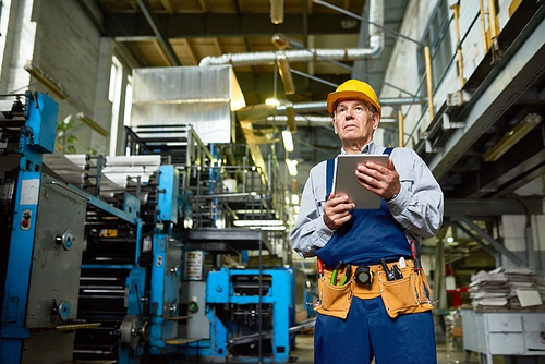 Low angle view of experienced engineer in protective helmet holding digital tablet in hands while wrapped up in work at manufacturing plant