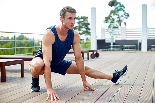 Handsome young sportsman doing side lunge while having training on spacious wooden terrace, picturesque view on background