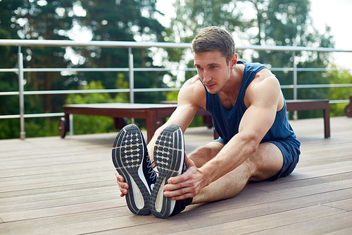 Confident young sportsman sitting on wooden floor of spacious terrace and doing stretching exercises before intensive workout, picturesque view on background