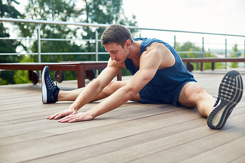 Young sporty man with closed eyes doing stretching exercise and enjoying fresh air while sitting on wooden floor of spacious terrace, picturesque view on background