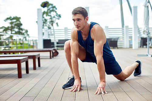 Portrait of muscular sportsman working out on wooden terrace outdoors stretching legs during warm up
