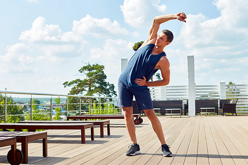 Active young man in sportswear enjoying breathtaking view while stretching arms before outdoor training at spacious terrace