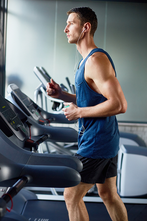 Profile view of young sporty man in headphones doing cardio exercise on treadmill, interior of modern gym on background