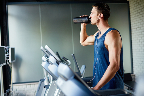 Concentrated sportsman in headphones refreshing himself with water while running on treadmill at gym, profile view