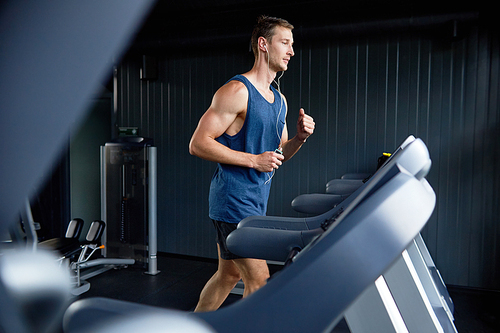 Full length portrait of muscular sportsman listening to music in headphones and using treadmill while wrapped up in training at modern gym, profile view