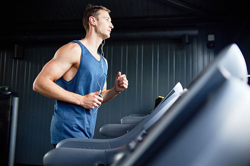 Side view portrait of muscular young man running on treadmill in modern gym during cardio workout, copy space