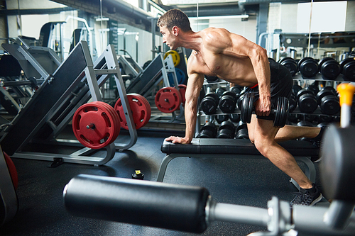 Side view portrait of handsome muscular man with bare chest working out with dumbbells on bench in modern gym, copy space