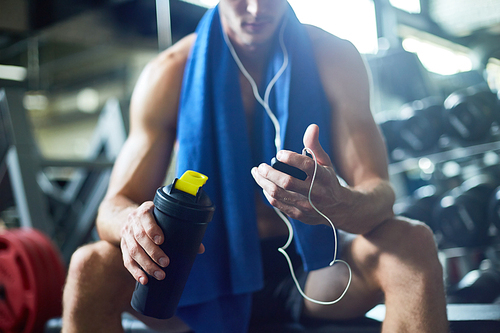Taking short break from intensive workout: young sporty man listening to music in headphones and holding bottle with water in hand while sitting on bench at modern gym