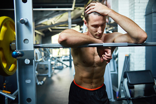 Exhausted young athlete with bare muscular torso leaning on barbell while taking short break from intensive workout at spacious gym, portrait shot
