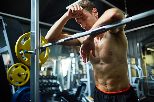 Waist-up portrait of muscular young man with closed eyes leaning on barbell while taking break from intensive workout at spacious gym